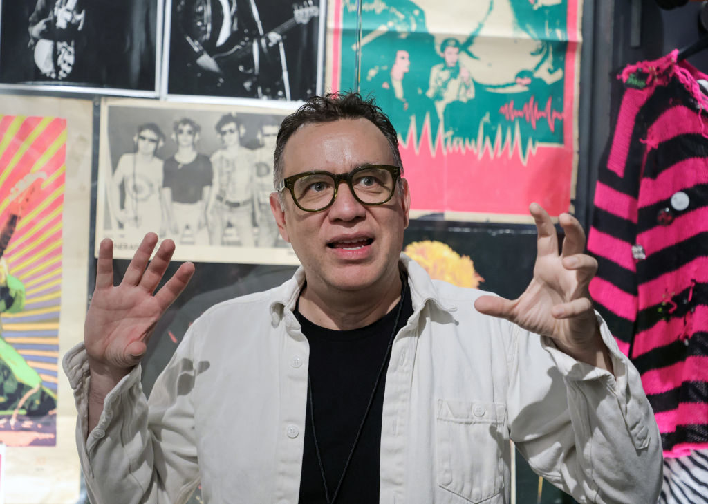 A person stands with hands raised in front of punk rock posters, wearing glasses and a white shirt over a black top