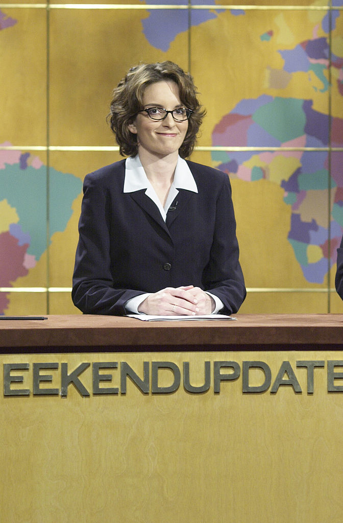 A woman in a suit sits at a news desk with &quot;Weekend Update&quot; visible. A colorful world map is in the background