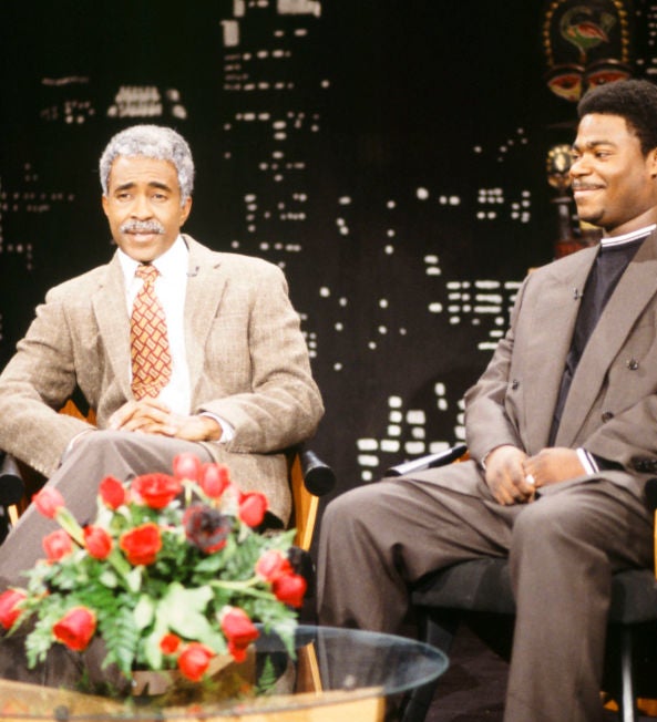 Two men sit on a TV set with a city backdrop. One wears a suit with a tie; the other a casual suit with a shirt. A flower arrangement is on the table