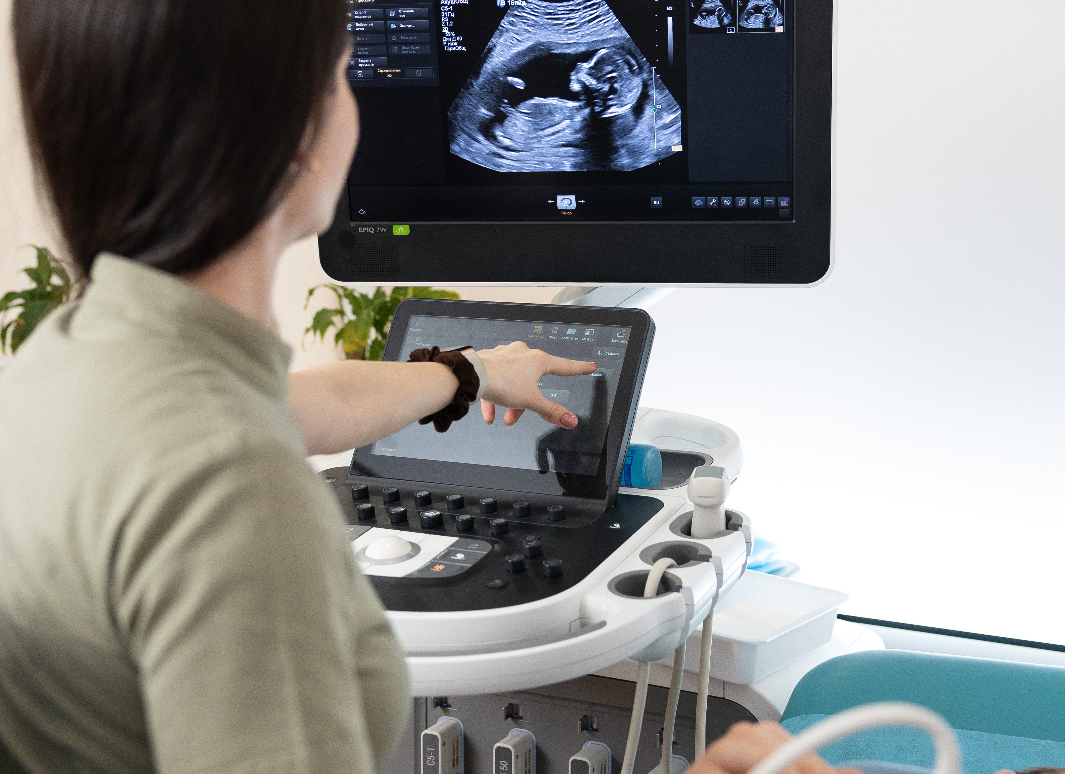 A technician performs an ultrasound on a pregnant person, pointing at the monitor displaying the baby&#x27;s image