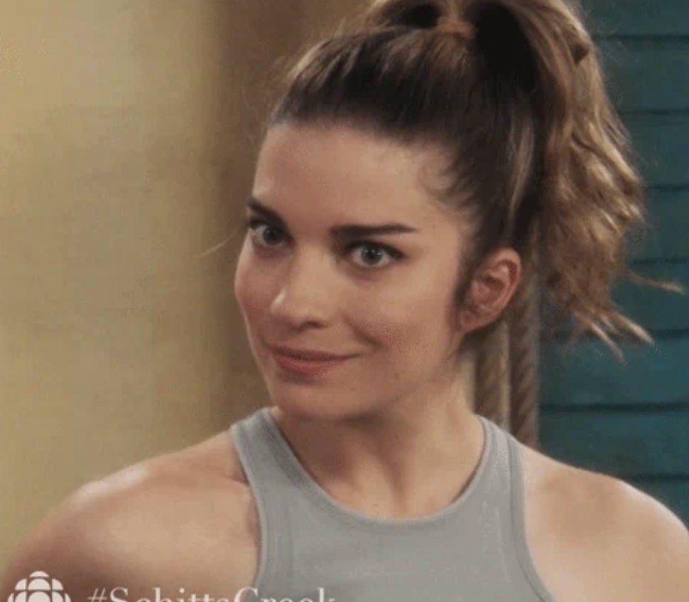 A woman in a high ponytail, wearing a sleeveless top, looks sideways with curiosity. Text: #SchittsCreek