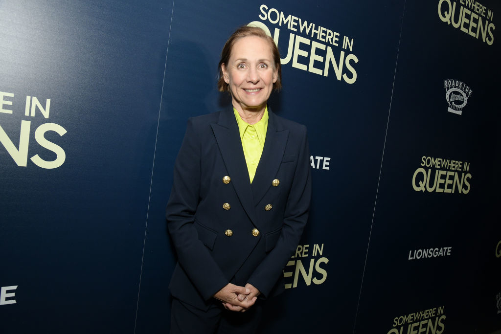 I don&#x27;t know who this person is. They are standing in front of a &quot;Somewhere in Queens&quot; backdrop, wearing a double-breasted suit and a bright shirt