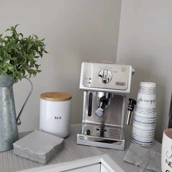 Espresso machine on a counter with a plant, tea canister, and stacked cups nearby