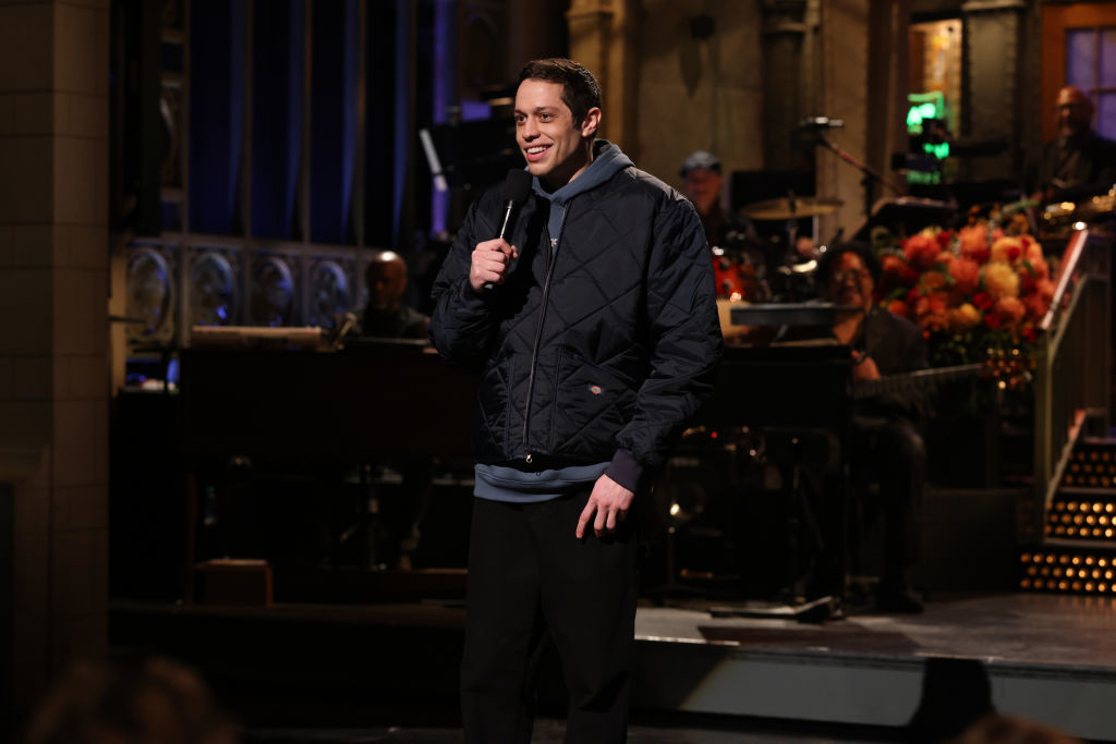 Comedian performing stand-up on a live TV show stage, wearing a casual quilted jacket over a hoodie