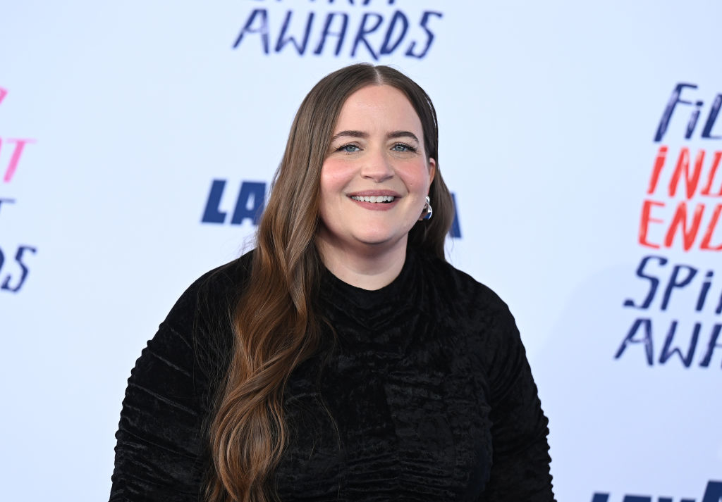 A person with long hair smiles, wearing a black outfit, at the Film Independent Spirit Awards backdrop