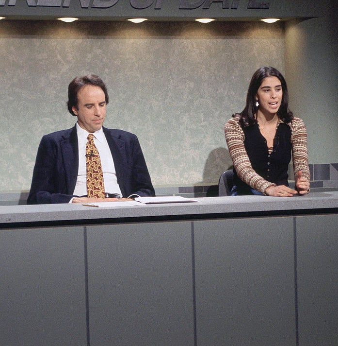 Two people are sitting at a news desk. The setting is a parody news segment titled &quot;Weekend Update.&quot;