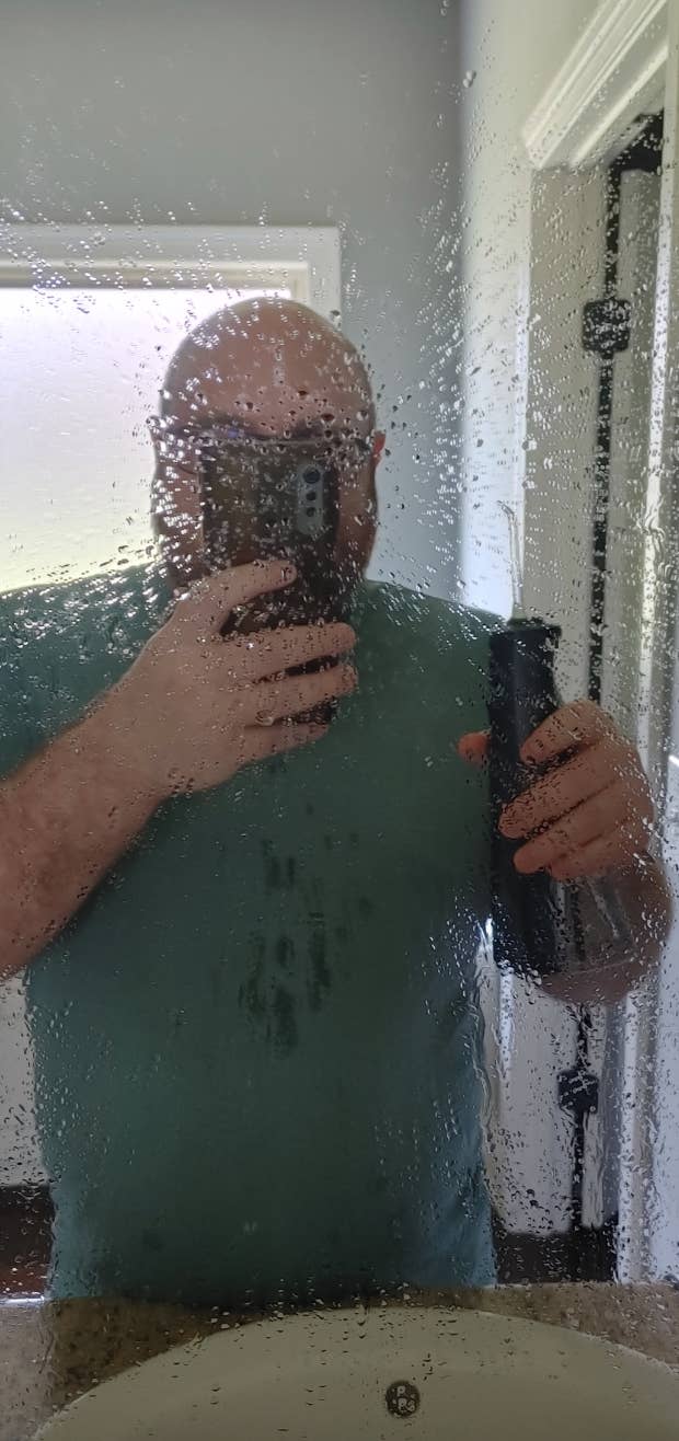 Man in a bathroom mirror taking a selfie with a phone, holding a water pick in the other hand