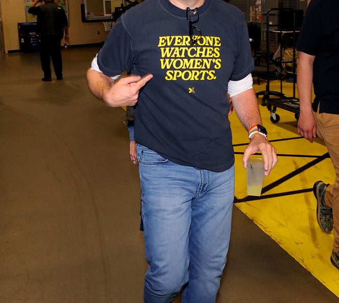 Man wearing a shirt with &quot;EVERYONE WATCHES WOMEN&#x27;S SPORTS&quot; text, jeans, and green shoes, walking down a hallway holding a drink cup