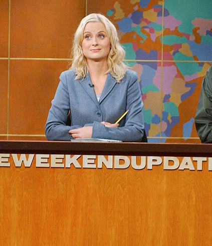 Tina Fey in a dark suit, Amy Poehler in a light suit, and a man in casual wear are at the &quot;Weekend Update&quot; desk on a news-style set with a world map