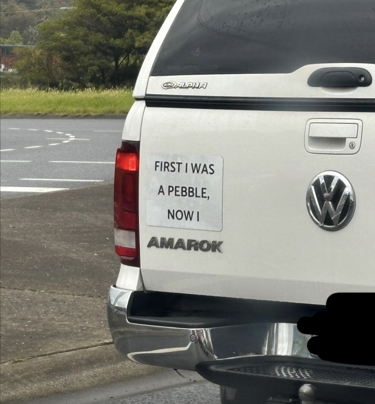 Volkswagen Amarok with a sign on the back reading, &quot;First I was a pebble, now I,&quot; followed by obscured text. Roadside background