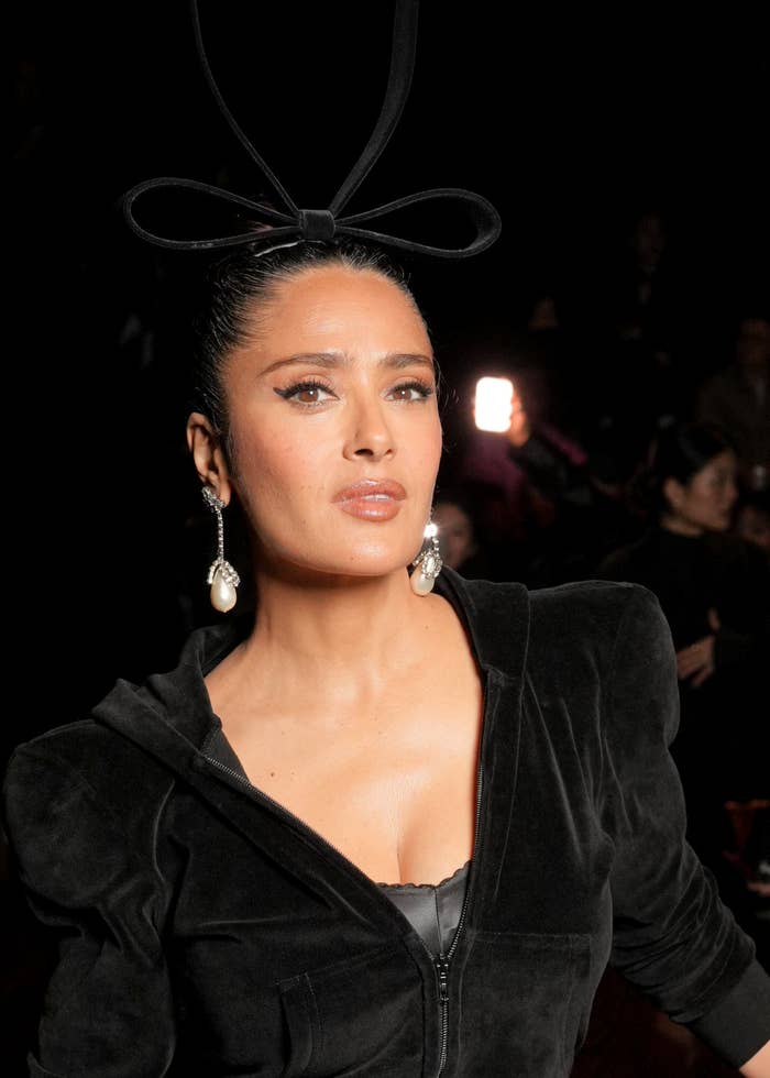 Salma Hayek con un elegante atuendo oscuro con un tocado de lazo único, posando sobre un fondo negro
