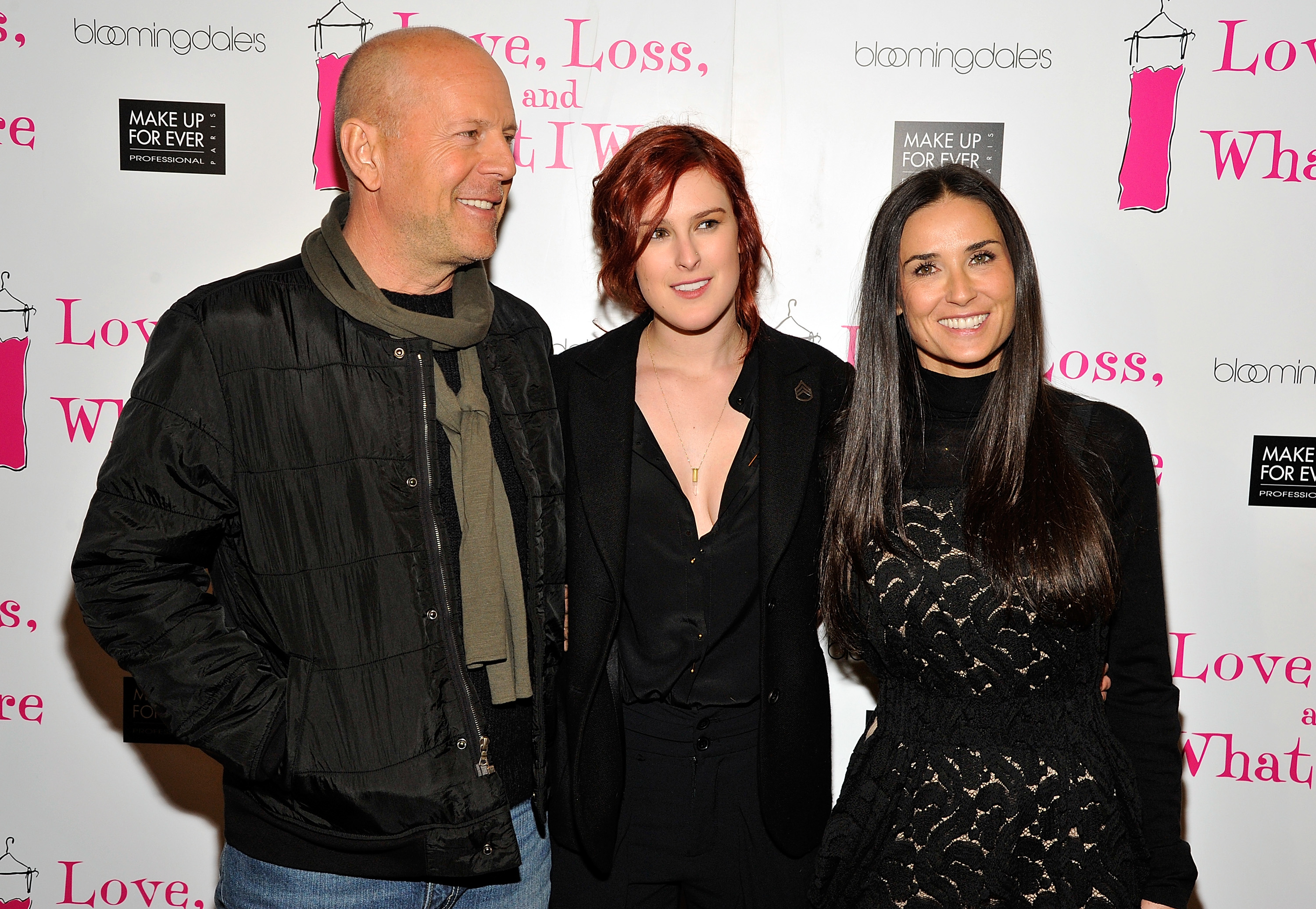 Three people smiling at an event: a person in a jacket and scarf, a person in a black suit, and a person in a black dress