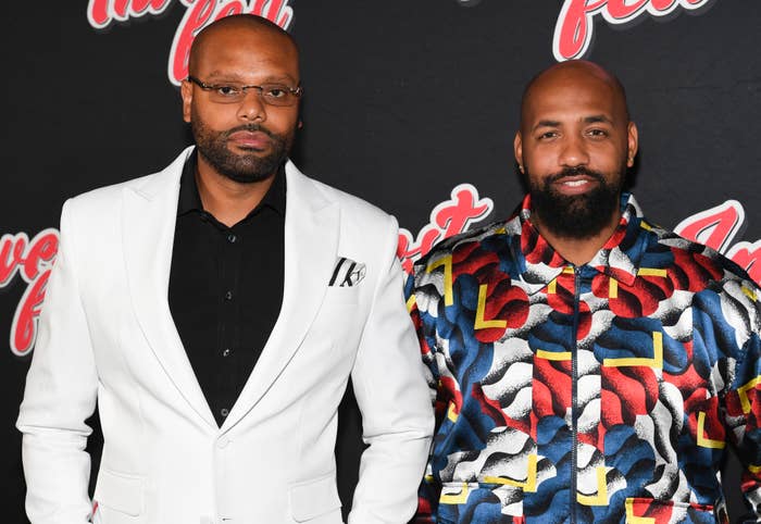 Two men on a red carpet. One wears a white blazer, the other a vibrant patterned shirt