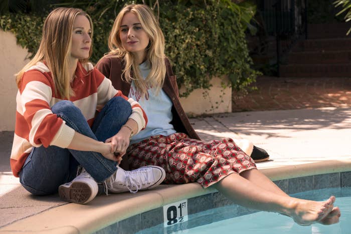 Two women, including Justine Lupe, sitting by a pool, wearing casual outfits with long sleeves
