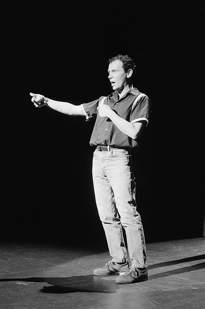 Man on stage performing in casual jeans and shirt, making a pointing gesture