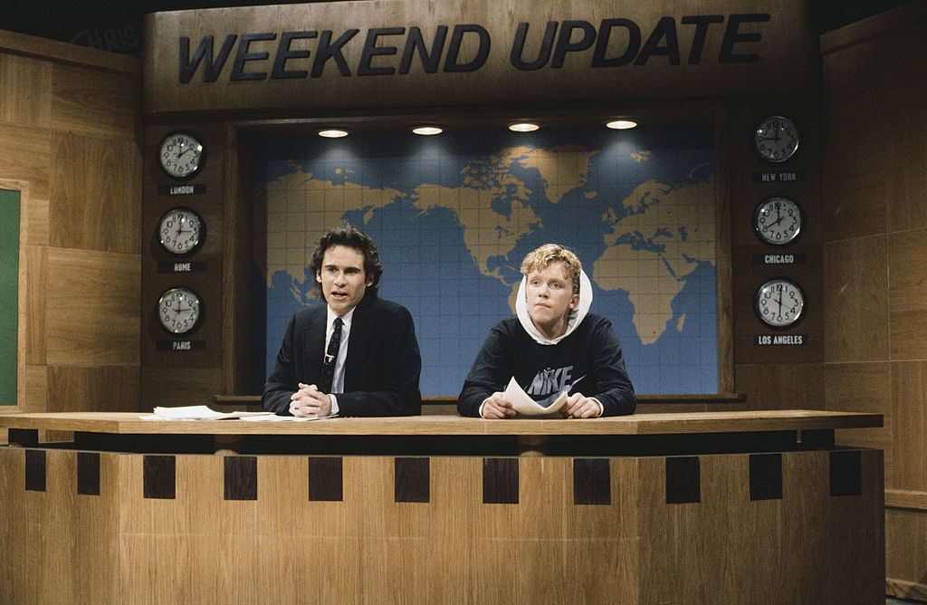 Two men on a news parody set. Left wears a suit; right is in a hoodie with a Nike logo. Clocks and world map on the wall