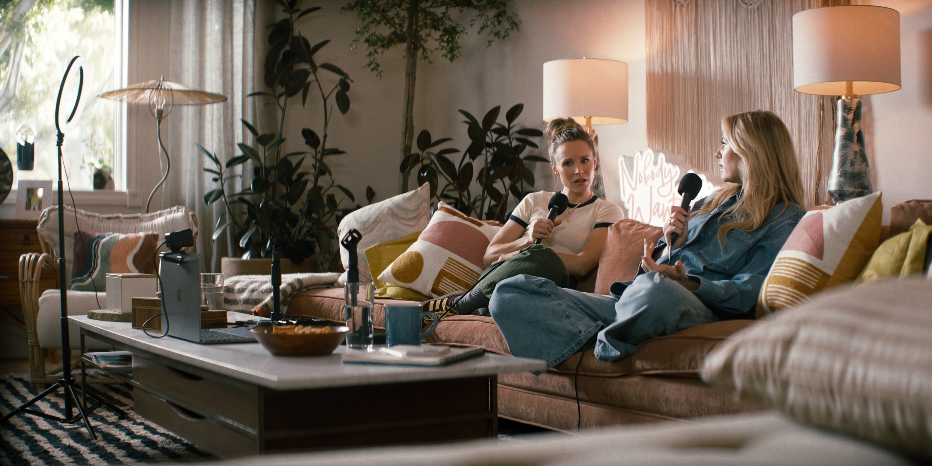 Two people sitting on a sofa with microphones, recording a podcast in a cozy living room setting with plants and soft lighting