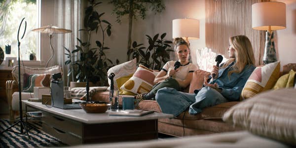 Two people sitting on a sofa with microphones, recording a podcast in a cozy living room setting with plants and soft lighting