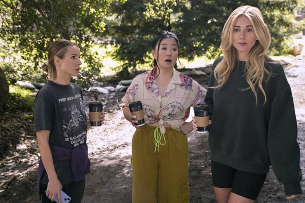 Three women outdoors with coffee cups, dressed casually in t-shirts and shorts, standing in a wooded area