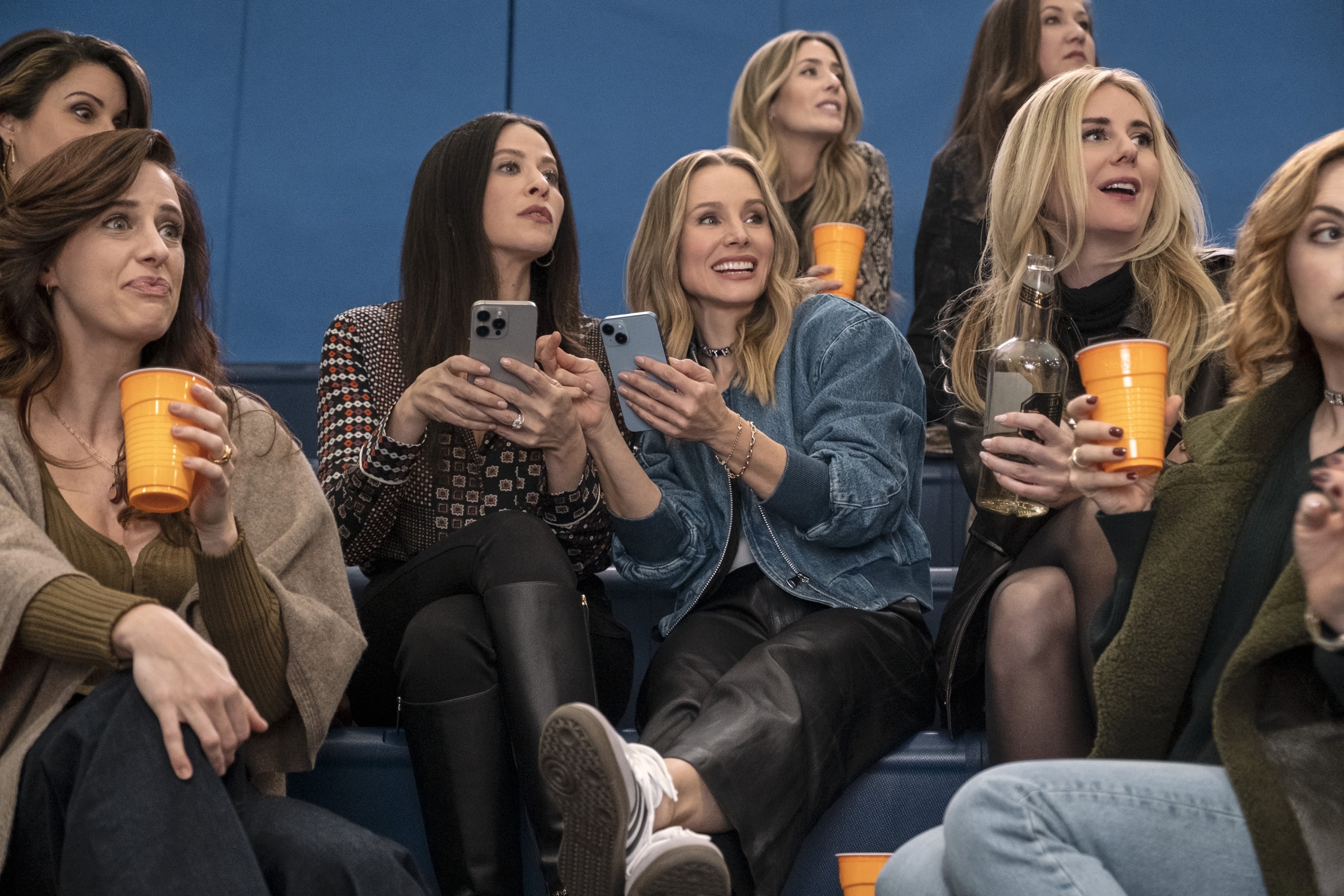 A group of women sit on bleachers holding drinks, casually dressed in jeans and jackets, watching an event