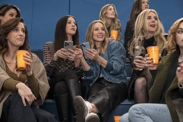 A group of women sit on bleachers holding drinks, casually dressed in jeans and jackets, watching an event
