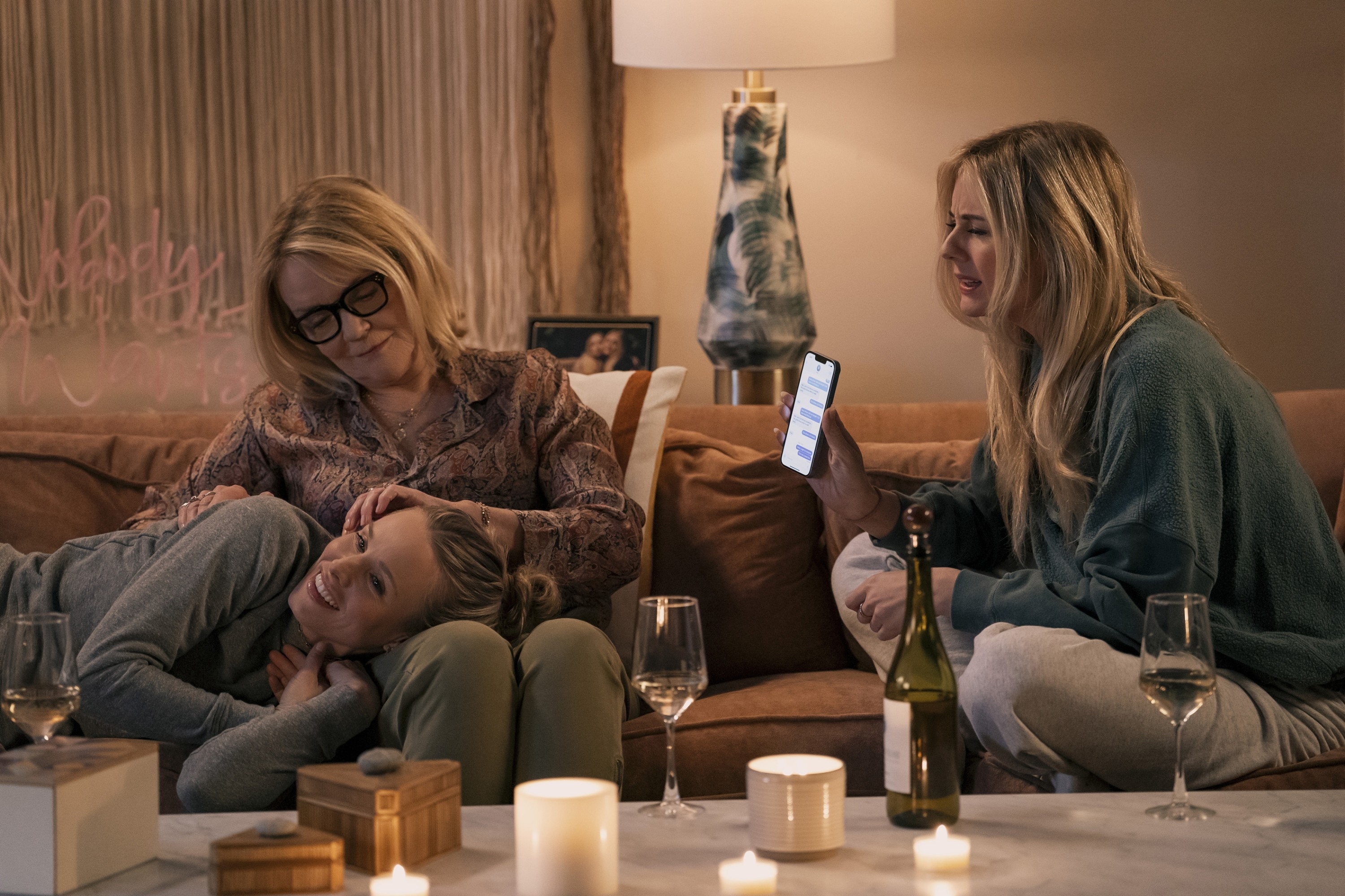 Three people relax on a couch with wine glasses and candles on a table nearby. One holds a phone displaying a message