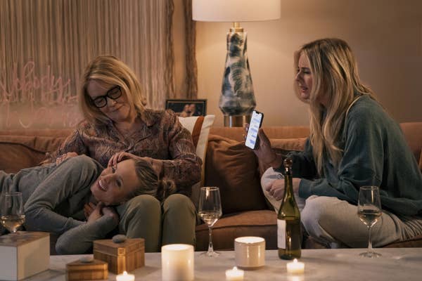 Three people relax on a couch with wine glasses and candles on a table nearby. One holds a phone displaying a message