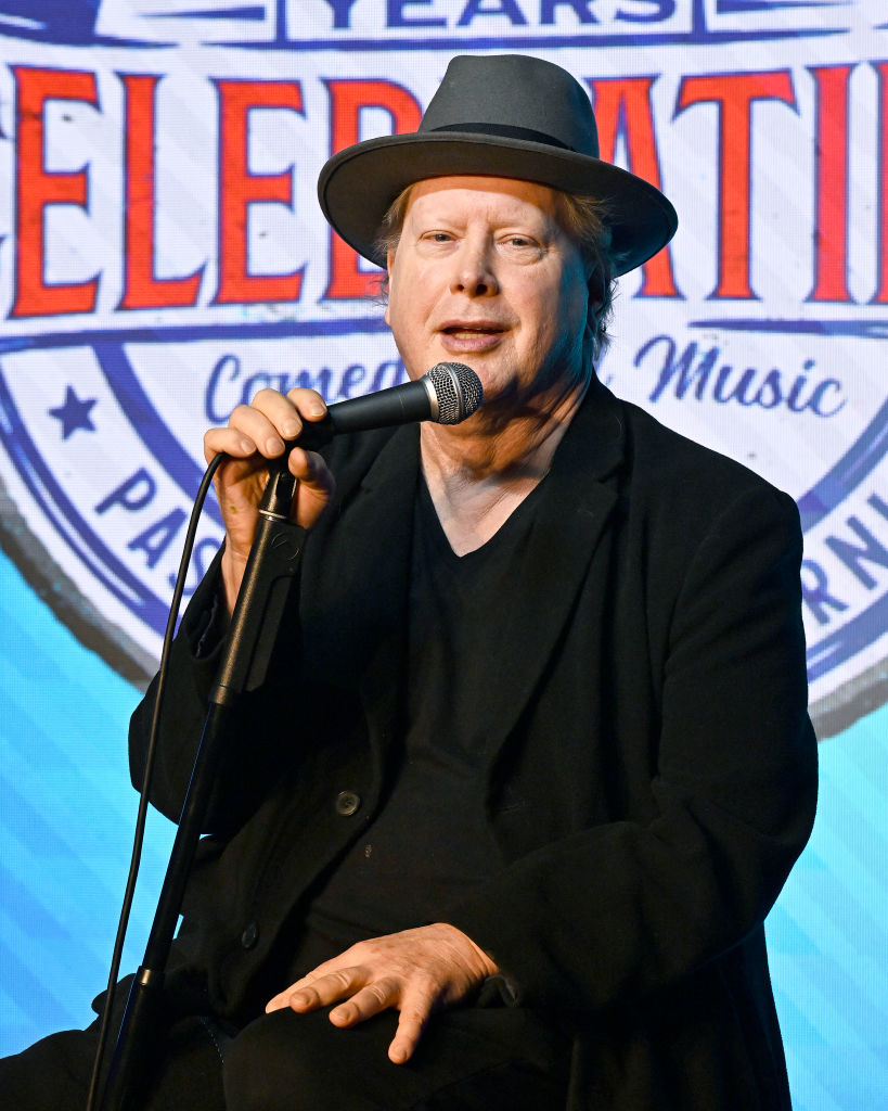 A person wearing a black suit and hat speaks into a microphone at an event with a celebratory banner in the background