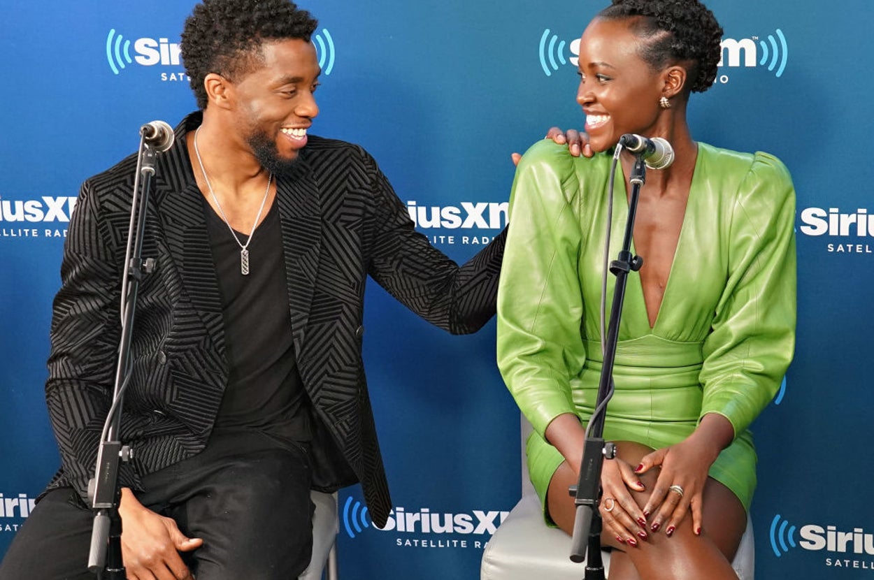 Two people seated, smiling at each other; one in a striped suit and the other in a green outfit, in front of a SiriusXM backdrop