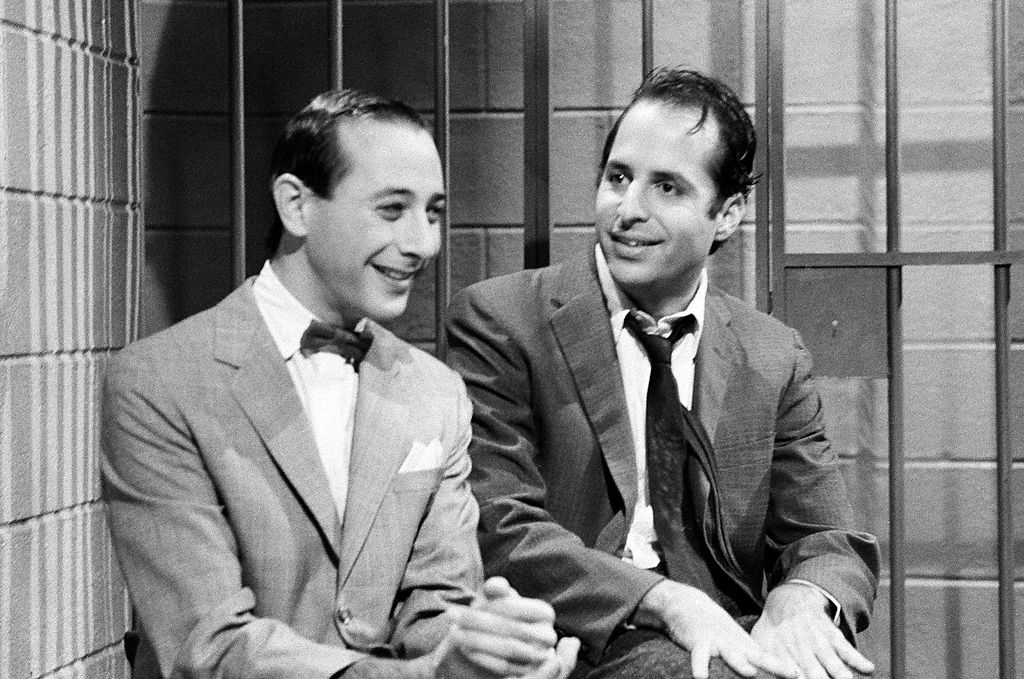 Two men in suits sit together, smiling, in front of a barred backdrop