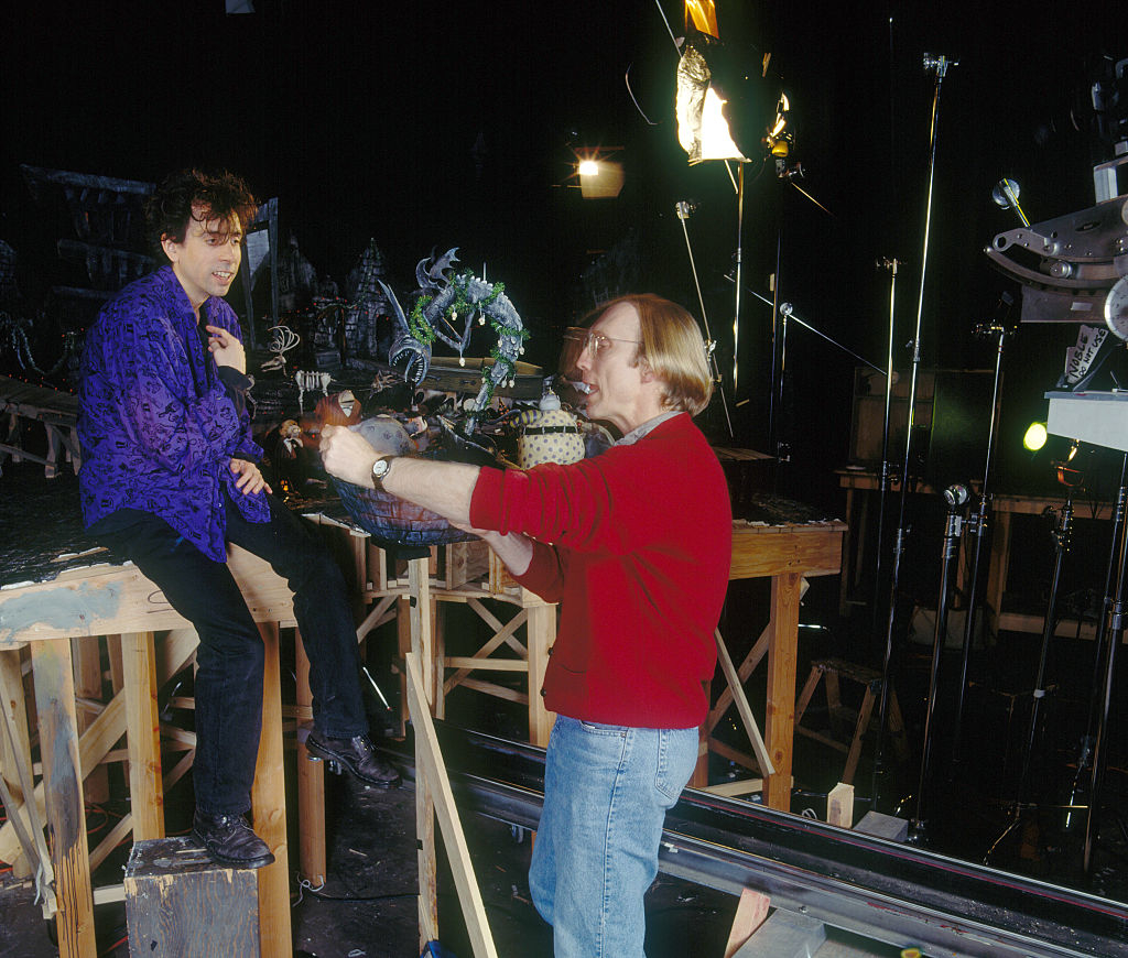 Two people on a film set, one sitting and the other holding a detailed puppet. The scene is filled with set elements and lighting equipment