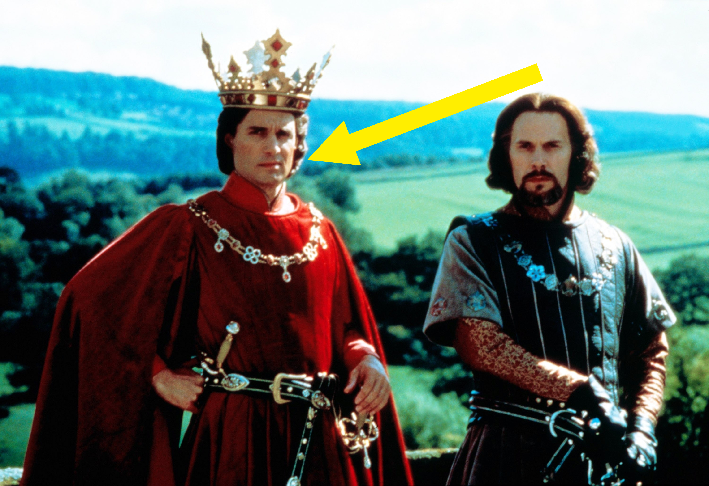 Two men in medieval attire standing outdoors; one wears a crown and red robe, the other has chain armor and a dark tunic