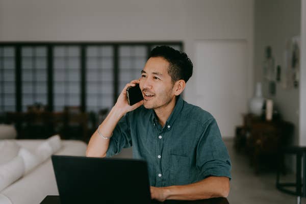 A person in a casual shirt sits at a table, smiling while talking on the phone with a laptop nearby