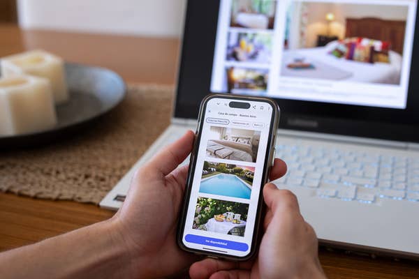 Person browsing vacation home rentals on a smartphone with a laptop open in the background, showing similar listings
