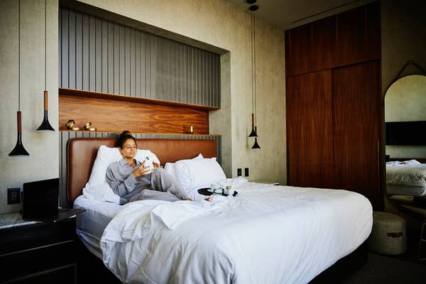 A person relaxing in a hotel room, sitting on a bed with breakfast on a tray, holding a phone and a cup. Cozy, modern interior design