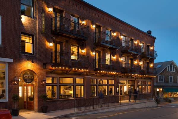 A charming brick hotel with glowing lights, featuring balconies and a street view at dusk. No people are visible in the image