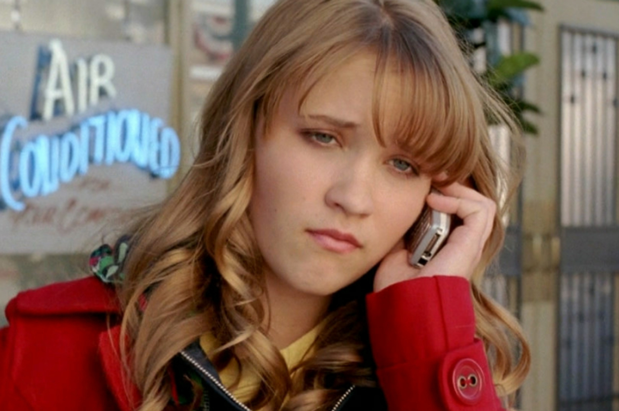 A person with wavy hair and a red coat listens on a phone, looking thoughtful. A neon sign in the background reads "Air Conditioned."