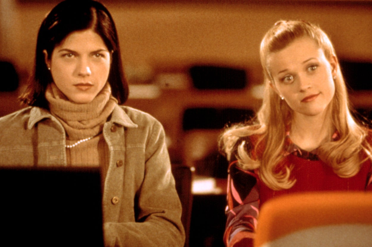 Two women seated indoors, one with dark hair in a turtleneck, the other with blonde hair wearing a stylish outfit