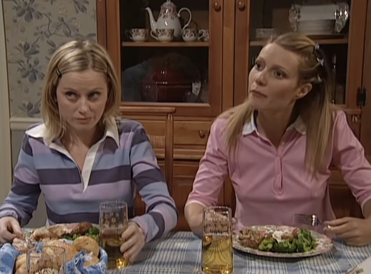 Two women seated at a dining table, one in a striped shirt, the other in a pink top. Comedic scene from a TV skit