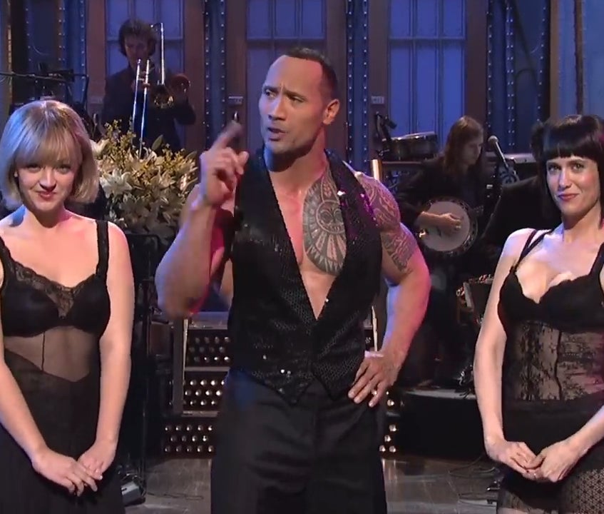 Three performers on a TV show stage: two women in black lace dresses and a man in a vest gesturing while speaking. Background features band members