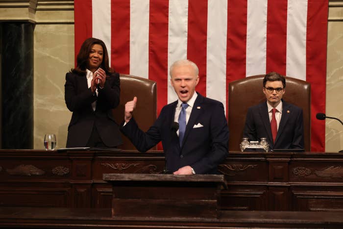 Mikey Day, as Biden in a suit, passionately speaks at a podium, while Punkie Johnson as Kamala Harris claps and, and Michael Longfellow, as Speaker Mike Johnson, observes on stage with an American flag backdrop during the 