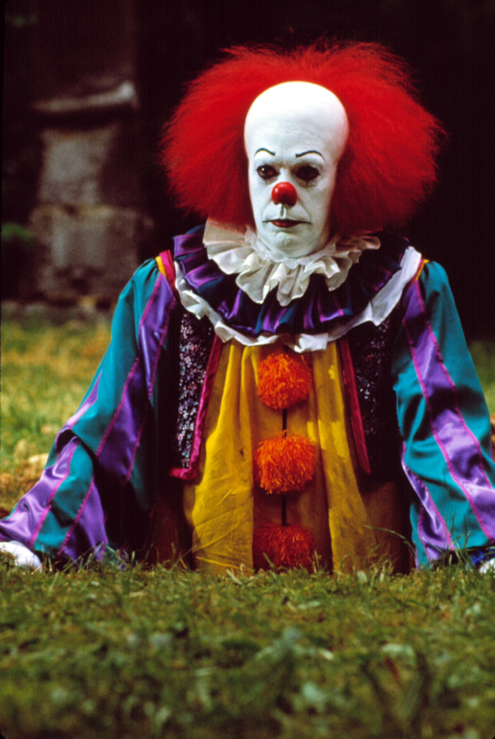 A creepy clown with red hair and colorful costume sits on the grass