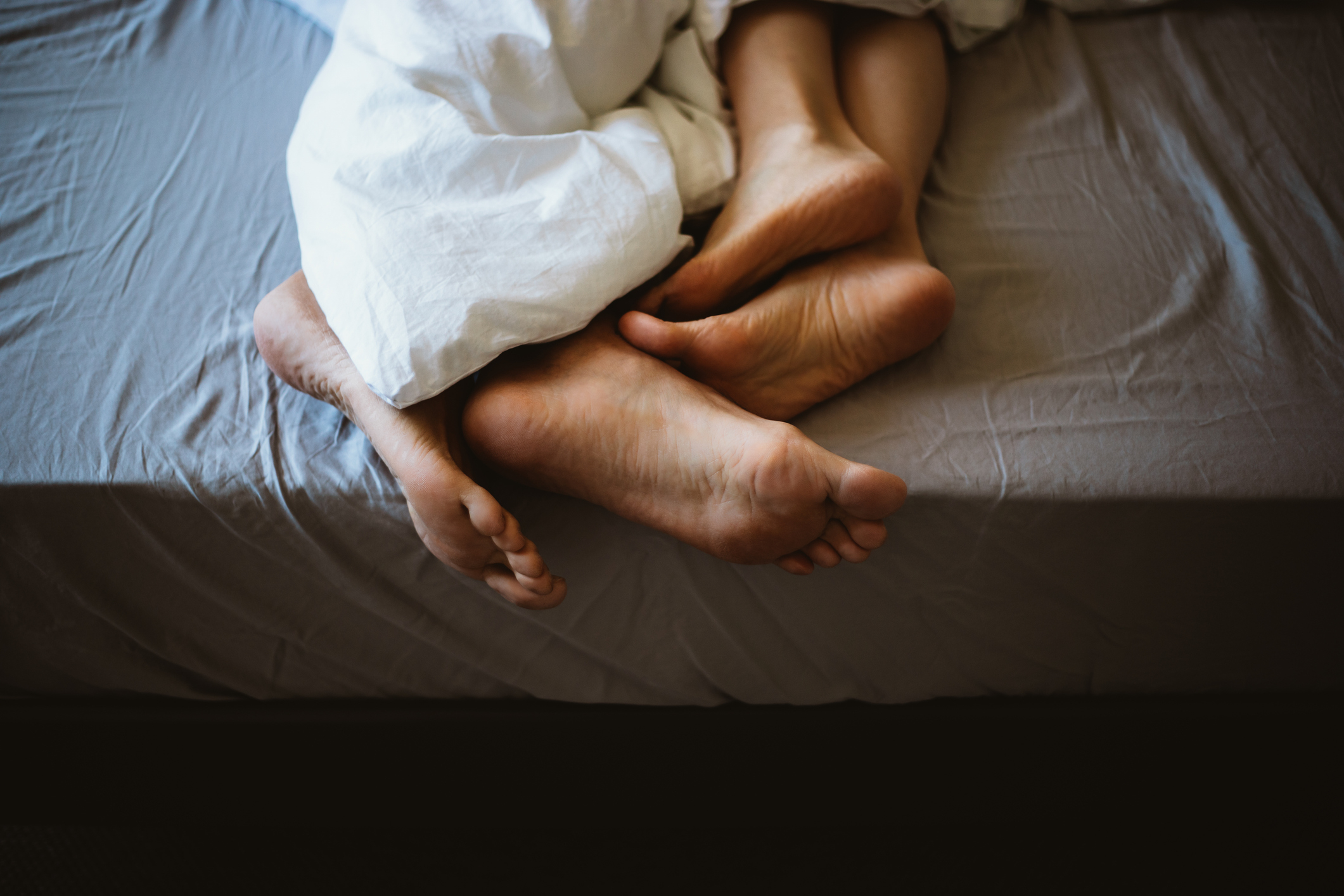 To par føtter viklet sammen under et teppe på en seng, noe som tyder på hygge og intimitet