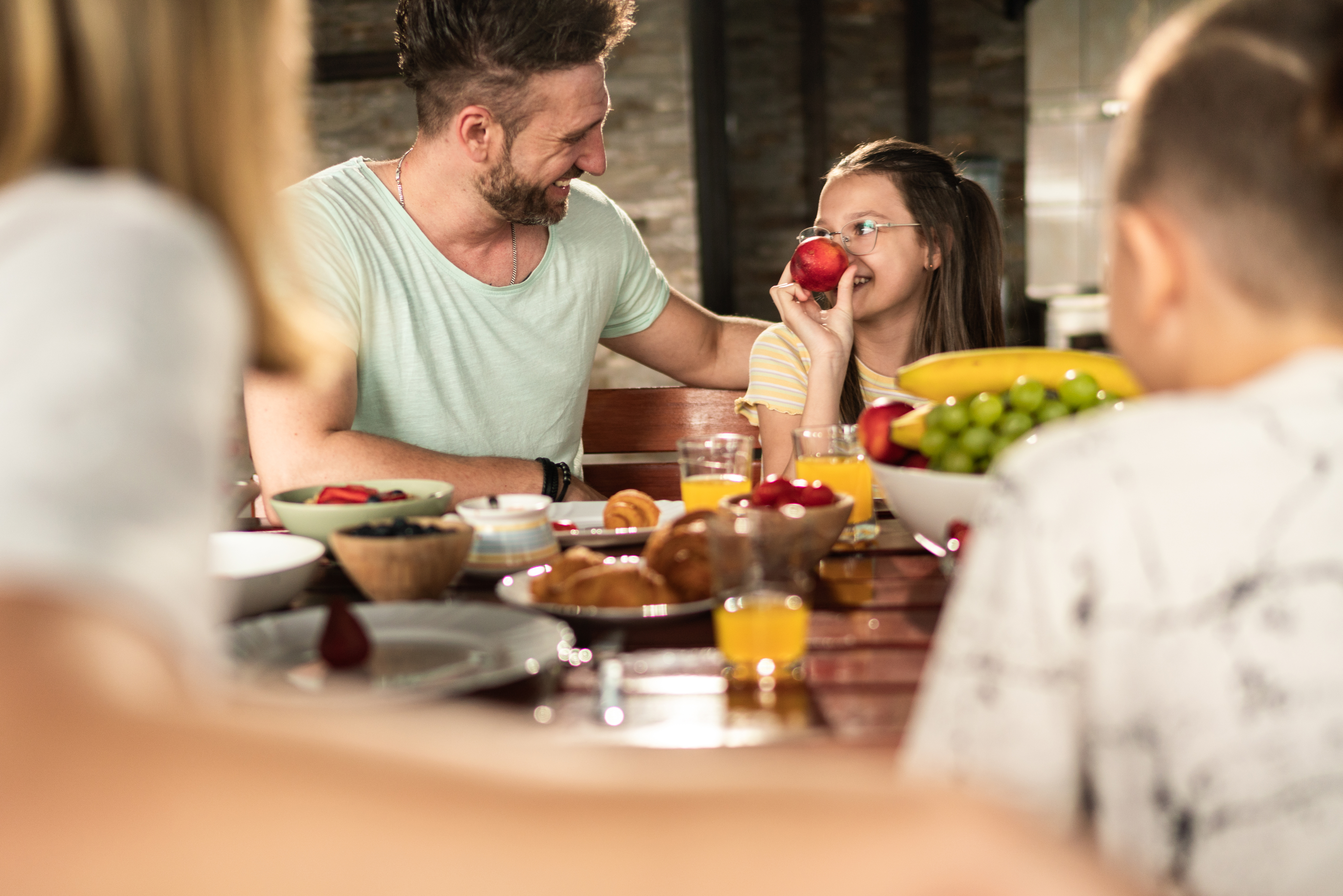 En familie sitter ved et bord og nyter et måltid. En mann smiler til en jente mens hun holder et eple for munnen, omgitt av frokostmat og frukt