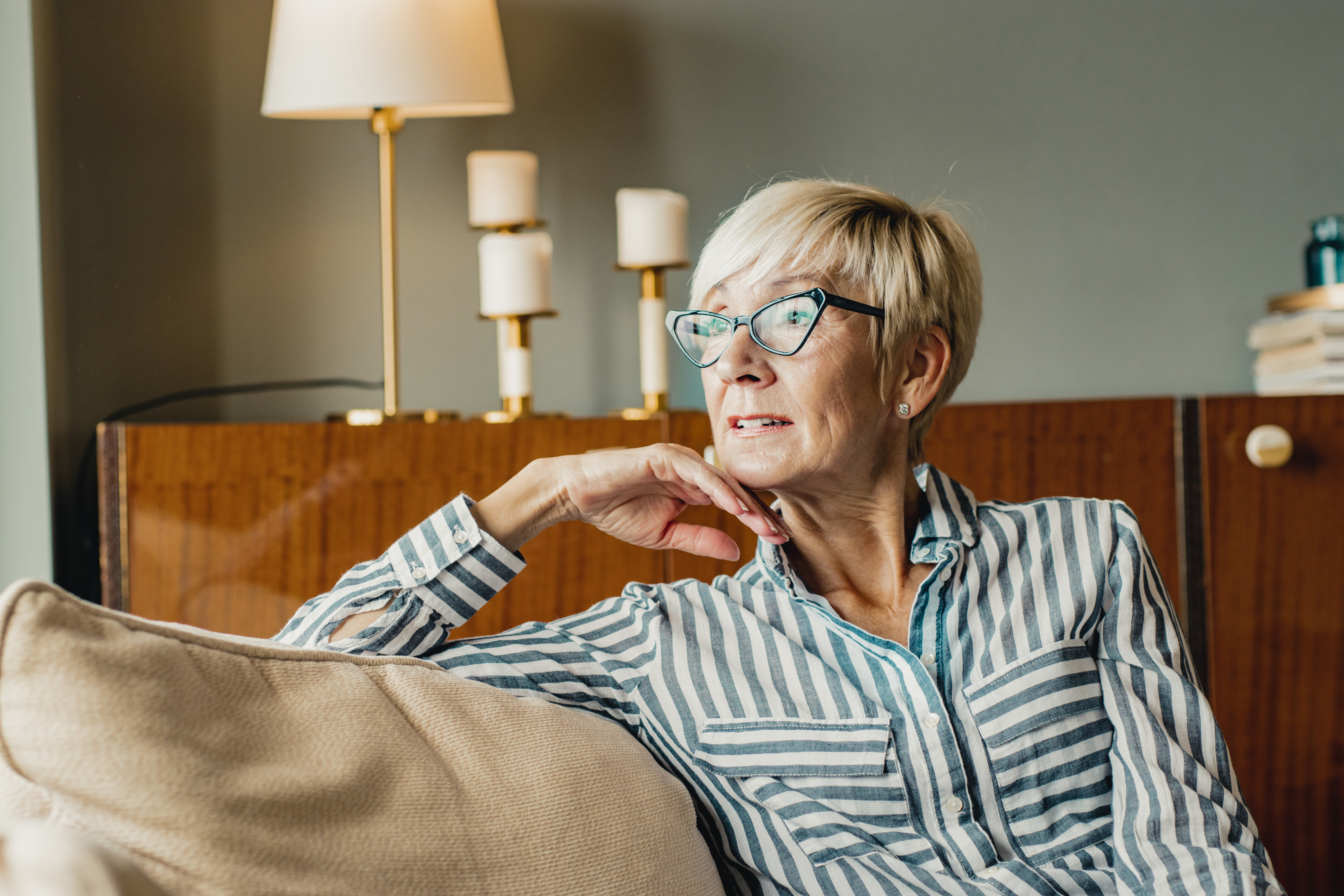 En eldre voksen med kort hår og briller slapper av i en sofa hjemme, iført en stripet skjorte. De fremstår som gjennomtenkte og innholdsrike