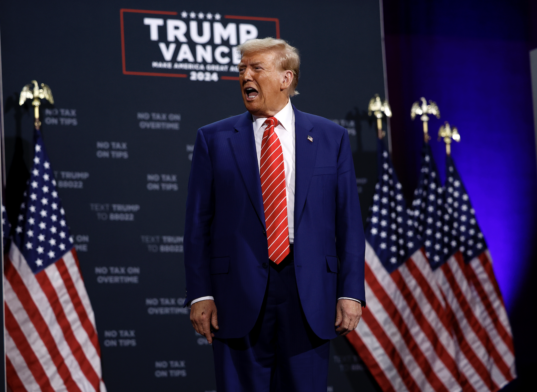 Trump i dress med stripet slips står på scenen med amerikanske flagg og et kampanjeskilt i bakgrunnen
