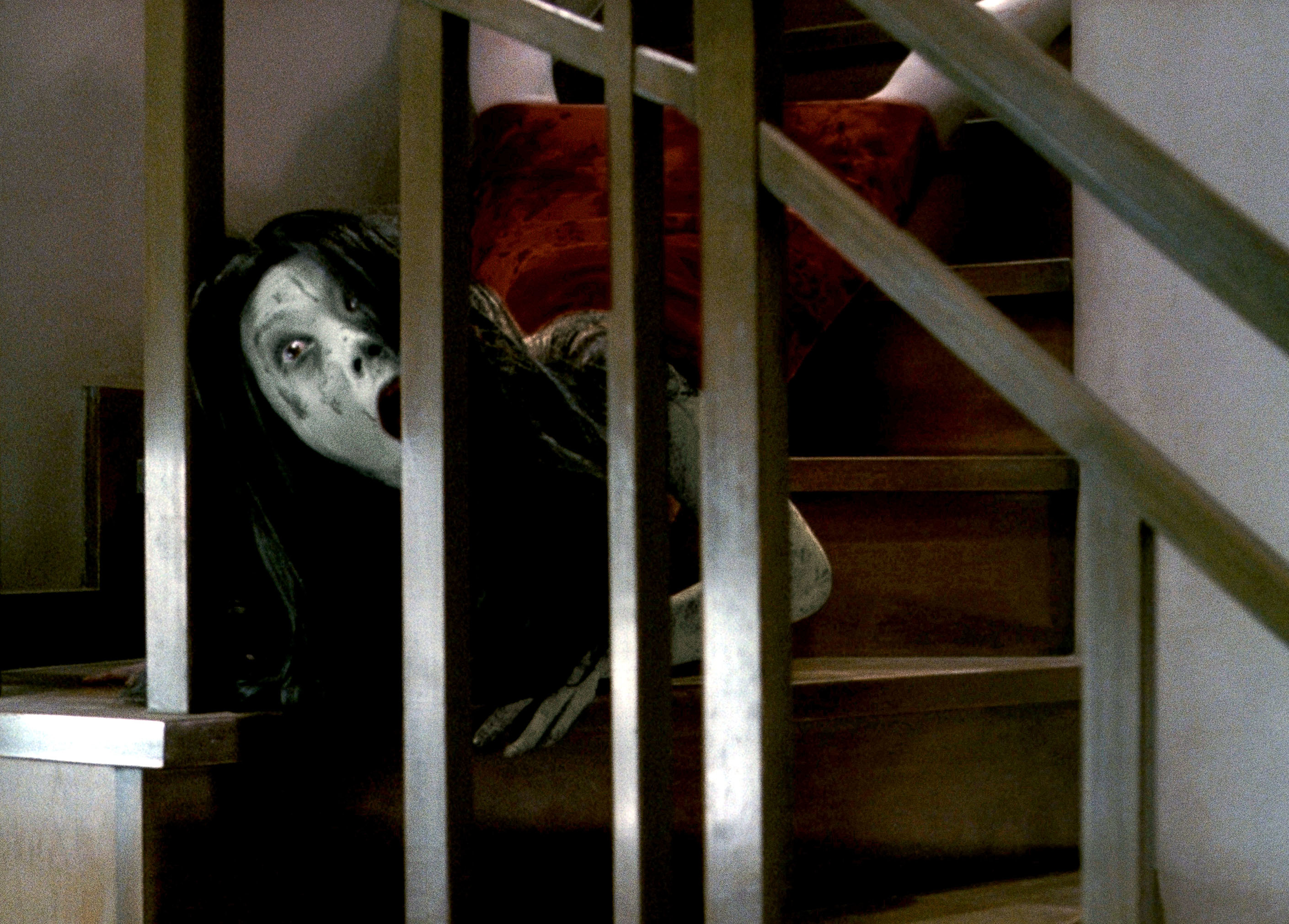 A ghostly figure with long hair and haunting expression crawls down a wooden staircase, emerging between the railings