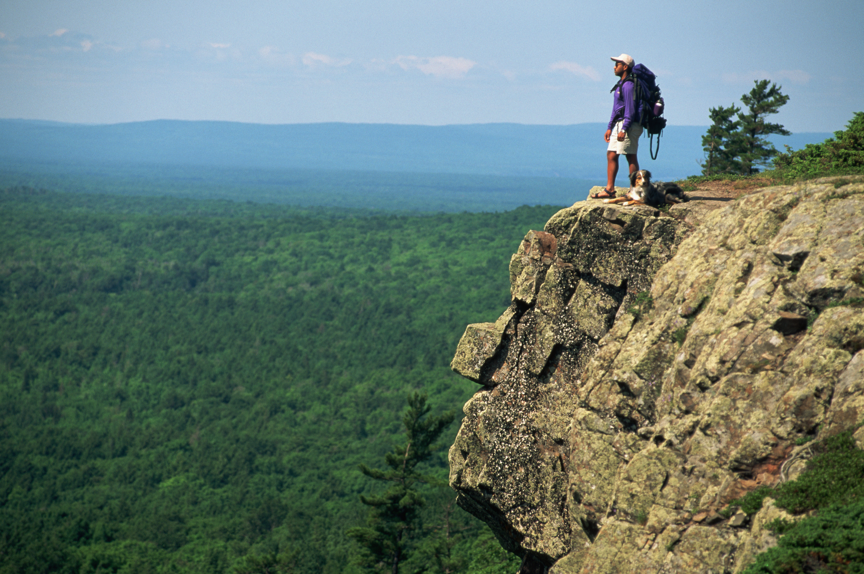 Person stands on cliff edge overlooking vast forest landscape with a backpack