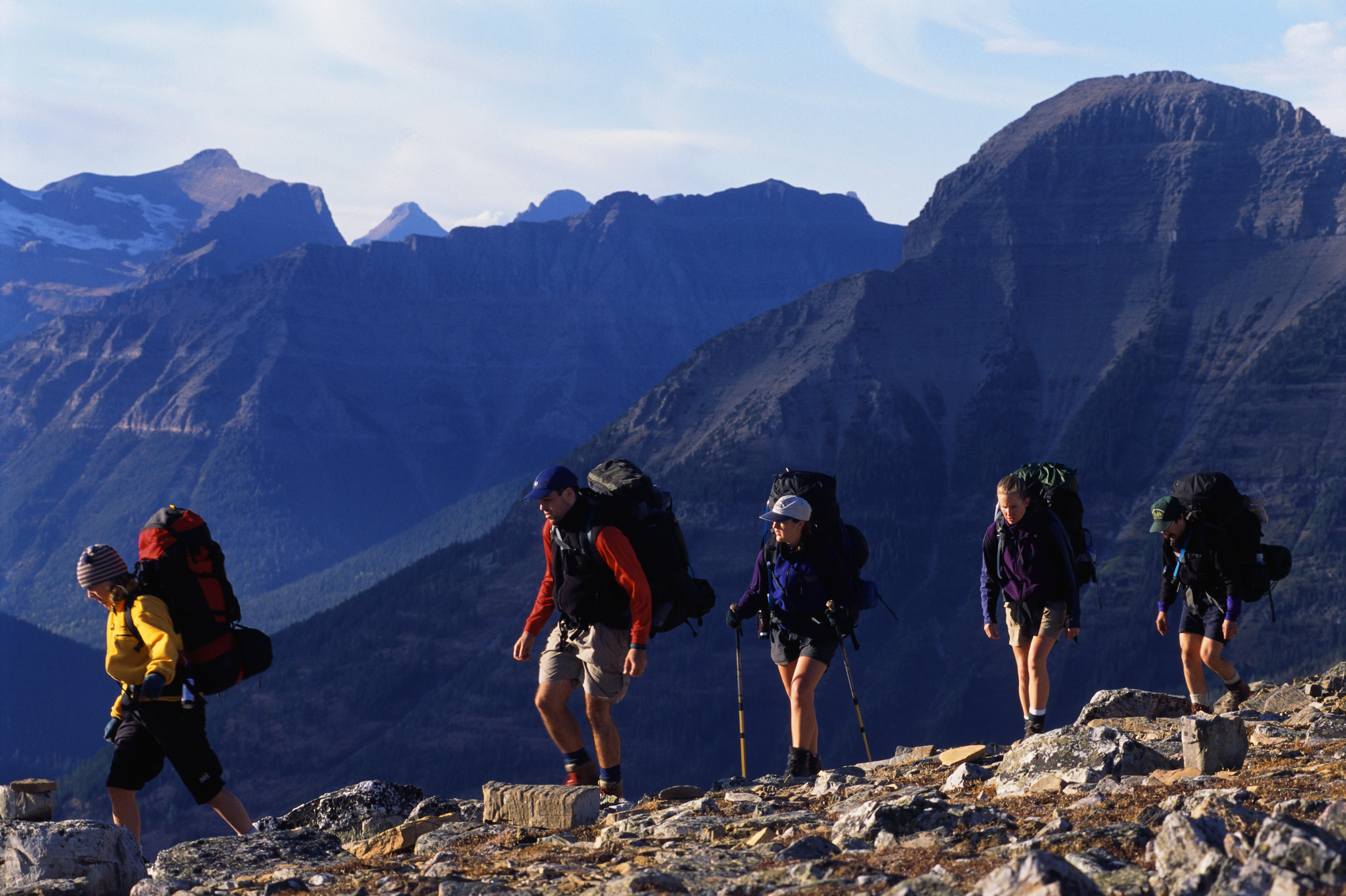 Five hikers with backpacks traverse a rocky mountain trail with tall peaks in the background under a clear sky