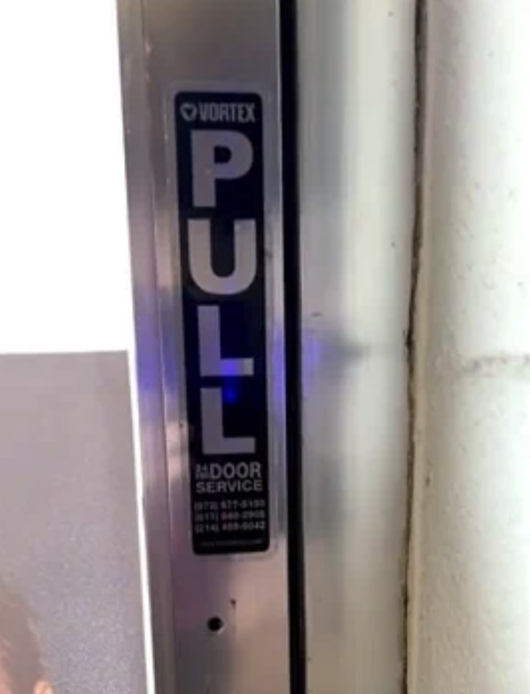 Metal door edge with a sign reading &quot;PULL&quot; and contact info for Vortex Door Service